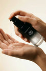 Hand holding a skincare bottle with dropper over another hand on a neutral background